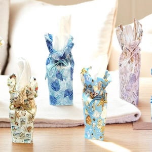 May include: A collection of fabric tissue box covers in various colors and patterns. The covers feature seashell and nautical designs in shades of blue, beige, and purple. Each cover is tied at the top, with white tissues visible.