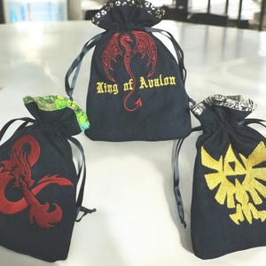 May include: Three black velvet drawstring bags with embroidered designs. The first bag has a red dragon with the text "King of Avalon". The second bag has a red Dungeons and Dragons symbol. The third bag has a gold Triforce symbol.