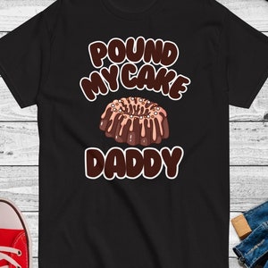 Puede incluir: Camiseta negra con el texto "POUND MY CAKE DADDY" en marrón y blanco, con un gráfico de un bizcocho. La camiseta se muestra sobre una superficie de madera clara. También se ven gafas de sol y una zapatilla roja.