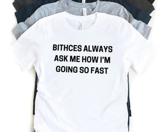 Puede incluir: Camiseta blanca con texto negro que dice "Bitches always ask me how I'm going so fast". La camiseta se muestra con varias otras camisetas de diferentes colores apiladas detrás de ella.