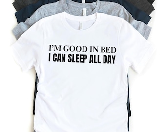 Puede incluir: Una camiseta blanca con texto negro que dice "I'm good in bed I can sleep all day". La camiseta se muestra con otras cinco camisetas de diferentes colores apiladas detrás de ella.