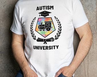 Puede incluir: Camiseta blanca con un diseño gráfico negro que dice "Autism University". El gráfico presenta un gorro de graduación, un tren de juguete y un diploma enrollado, todo dentro de una corona de hojas.