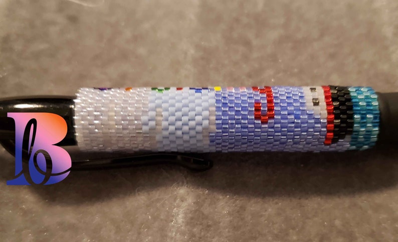 Puede incluir: Un bol&iacute;grafo negro con un dise&ntilde;o de cuentas en tonos de azul, blanco, rojo, negro y turquesa. Las cuentas est&aacute;n dispuestas en un patr&oacute;n que se asemeja a una bandera.