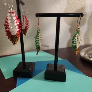 May include: A pair of beaded earrings on black display stands. The earrings are made of white, green, and brown beads and are shaped like leaves.