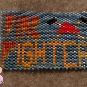 May include: Beaded bracelet with the words "FIRE FIGHTER" in orange and yellow beads on a blue background. A red and yellow beaded firefighter helmet with an axe is in the upper right corner.