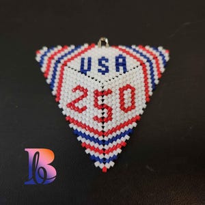 May include: A triangular beaded pendant in red, white, and blue. The pendant features the word "USA" in blue and the number "250" in red. The pendant has a silver-colored loop at the top for hanging.