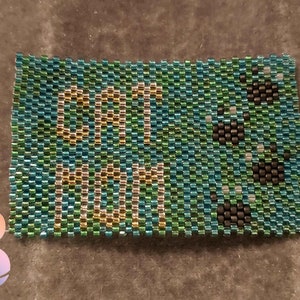 Könnte beinhalten: Ein Armband aus Perlen mit den Worten "Get Me Out" in goldenen Perlen auf einem türkisfarbenen, grünen und schwarzen Hintergrund. Das Armband hat ein Muster aus schwarzen Perlen.