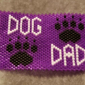 Op de afbeelding: Paarse kralenarmband met de woorden "DOG DAD" in witte kralen en twee zwarte poot afdrukken.