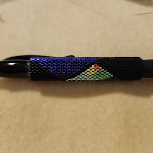 Puede incluir: Un bolígrafo negro con un diseño de cuentas en azul, negro, blanco, verde y naranja. Las cuentas están dispuestas en un patrón geométrico.