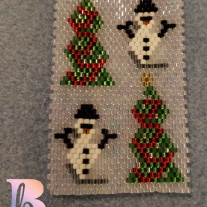 May include: A beaded Christmas ornament featuring two snowmen and two Christmas trees. The snowmen are white with black hats and black buttons. The trees are green with red and white stripes.