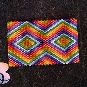 May include: A colorful beaded rectangle with a geometric pattern of diamonds in shades of red, orange, yellow, green, blue, and purple.