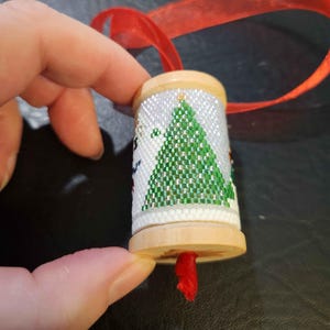May include: A wooden spool ornament with a white background and a green Christmas tree design. The ornament has a red ribbon attached to the top.