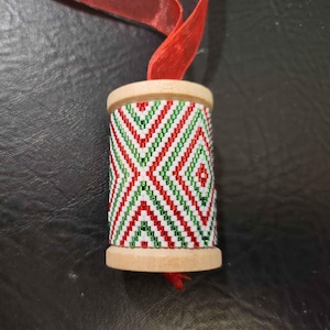 May include: A wooden spool ornament with a red, white and green geometric pattern. The spool is decorated with a red ribbon.