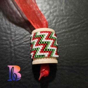 May include: A wooden spool with a red, white, and green beaded design. The spool is wrapped with a red ribbon.