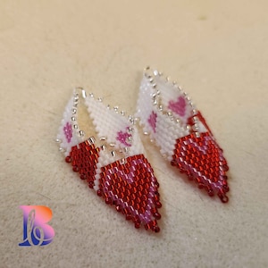 May include: A pair of dangle earrings with a white and red beaded design. The earrings are shaped like a teardrop and feature pink hearts.