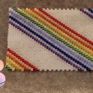 May include: A white beaded square with a diagonal rainbow pattern. The rainbow is made of red, orange, yellow, green, blue, indigo, and violet beads.