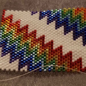 May include: A rainbow-colored beaded pattern with a white background. The beads are arranged in a zig-zag pattern, creating a geometric design.