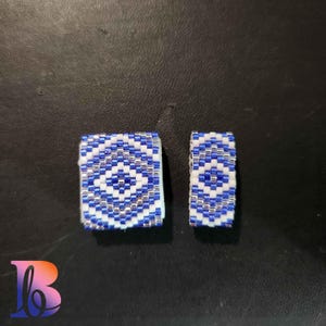May include: Two small, square, beaded earrings with a white and blue geometric pattern. The pattern features a central blue circle surrounded by white and silver beads.
