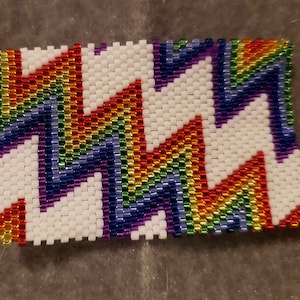 May include: A rainbow-colored beaded bracelet with a zig-zag pattern. The bracelet is made with small, colorful beads and is woven in a white background.