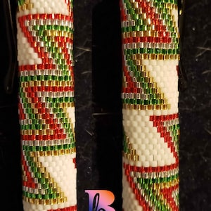 May include: Two beaded pen covers. The pen covers are white with a repeating pattern of red, green, and gold beads. The pen covers have a black clip. The pen covers are cylindrical.
