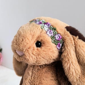 May include: A plush rabbit toy with soft tan fur, long floppy ears, and a small pink nose. The rabbit features a decorative headband across its forehead, embroidered with small purple and white flowers and green leaves on a lace-like trim.