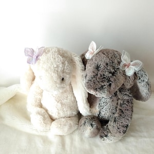 May include: Two plush bunny rabbits, one cream-colored and the other gray, sit side-by-side. Each bunny has a delicate butterfly accent on its head. The bunnies are made of soft, textured material and are positioned on a white, fluffy surface.