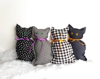 May include: Four stuffed fabric cat figures in a row. The first is black with white polka dots and a purple ribbon. The second is gray with black polka dots and a purple ribbon. The third is black and white gingham with an orange ribbon. The fourth is black with white polka dots and an orange ribbon.