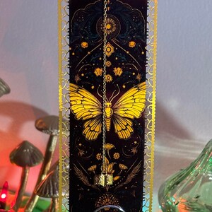 Puede incluir: Una decoración colgante con marco dorado, fondo negro y una mariposa amarilla con diseños florales. La decoración colgante tiene una cadena dorada con un pequeño amuleto dorado.