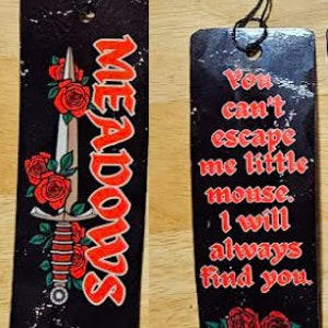 May include: Black bookmark with a red and white sword and red roses. The text "MEADOWS" is written in red on the bookmark. The other side of the bookmark has black text on a white background that reads "You can't escape me little mouse, I will always find you."