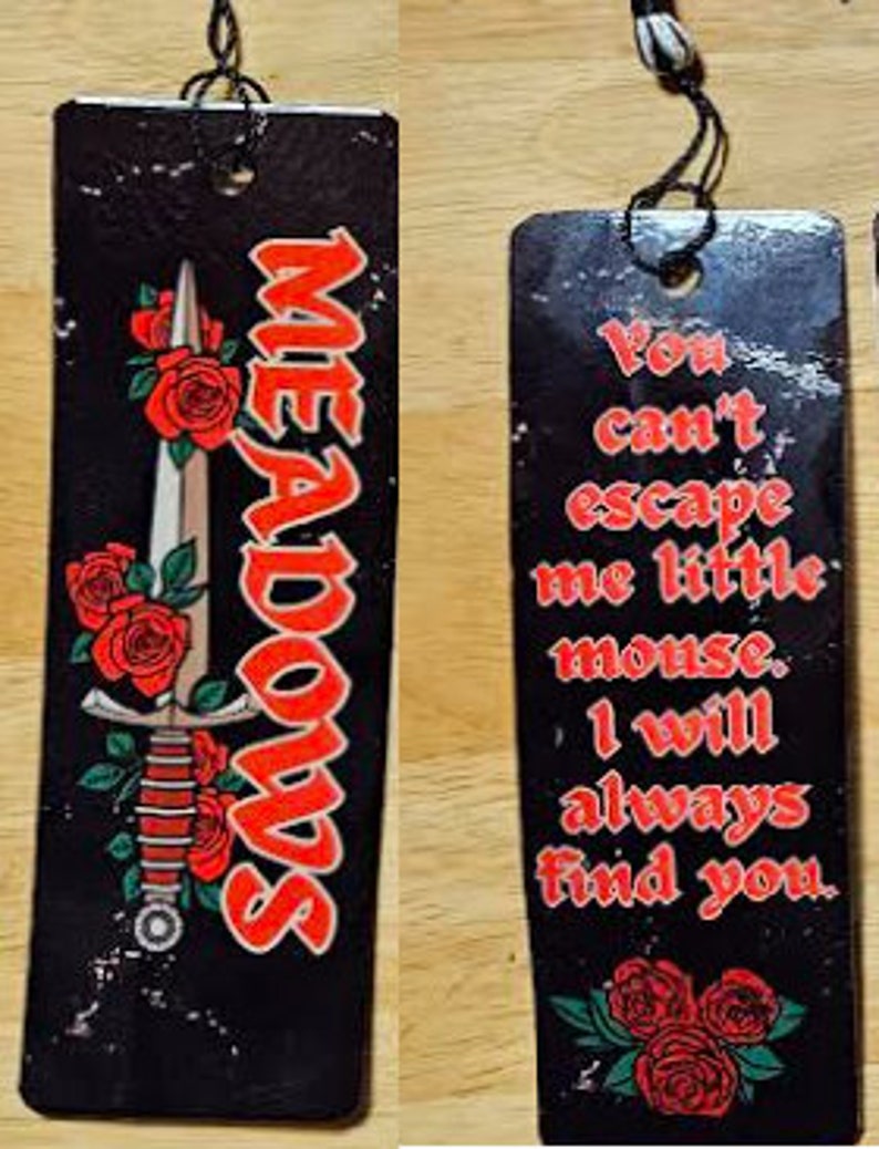 May include: Black bookmark with a red and white sword and red roses. The text "MEADOWS" is written in red on the bookmark. The other side of the bookmark has black text on a white background that reads "You can't escape me little mouse, I will always find you."