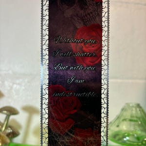 May include: A silver metal bookmark with a decorative border. The bookmark features a skull and roses with the text "Without you, I will shatter. But with you, I am indestructible."
