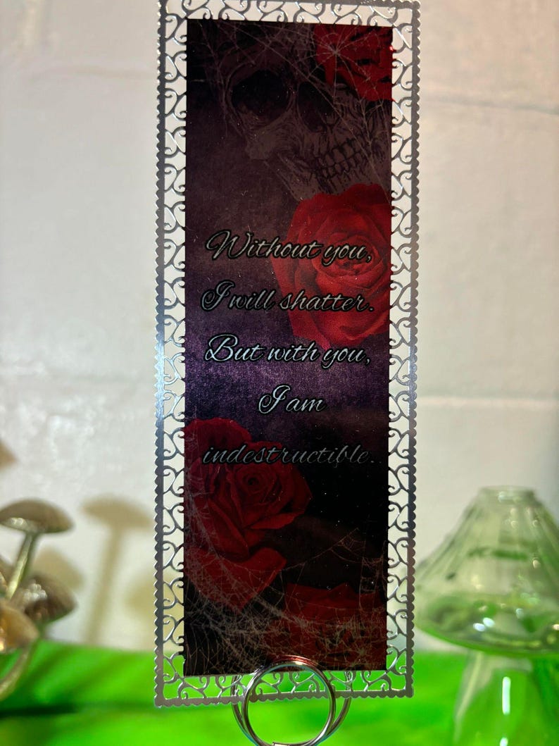 May include: A silver metal bookmark with a decorative border. The bookmark features a skull and roses with the text "Without you, I will shatter. But with you, I am indestructible."