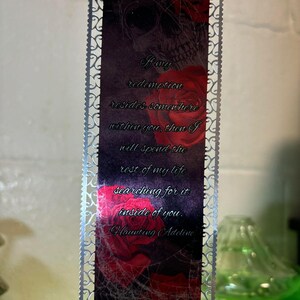 May include: A silver bookmark with a black and red design featuring a skull, roses, and spiderwebs. The bookmark has a quote that reads: "If my redemption resides somewhere within you, then I will spend the rest of my life searching for it inside of you." - Haunting Adeline
