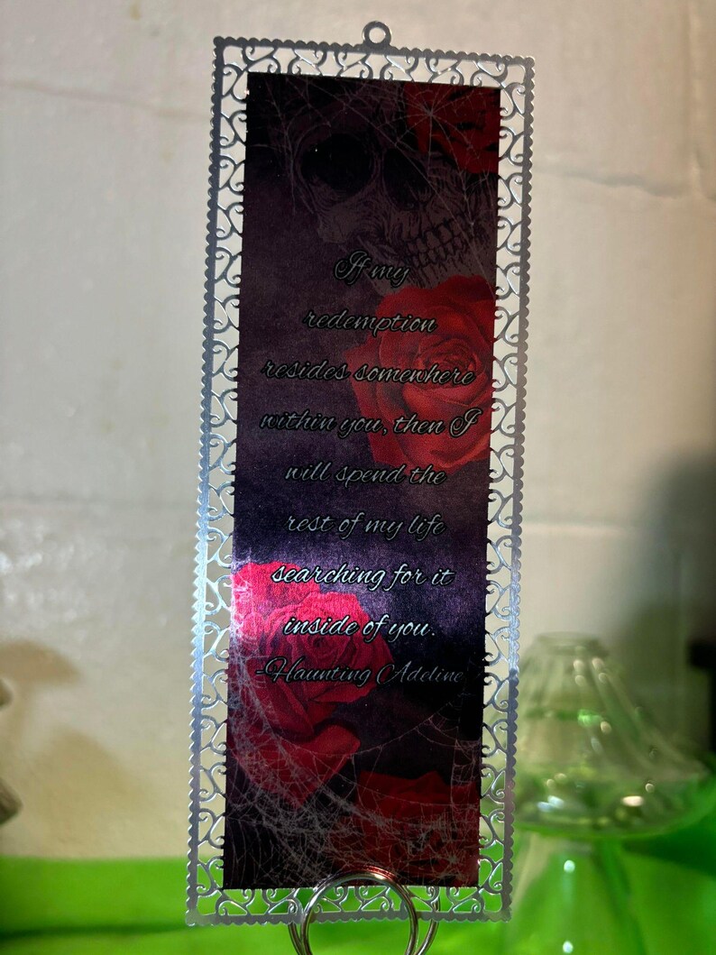 May include: A silver bookmark with a black and red design featuring a skull, roses, and spiderwebs. The bookmark has a quote that reads: "If my redemption resides somewhere within you, then I will spend the rest of my life searching for it inside of you." - Haunting Adeline