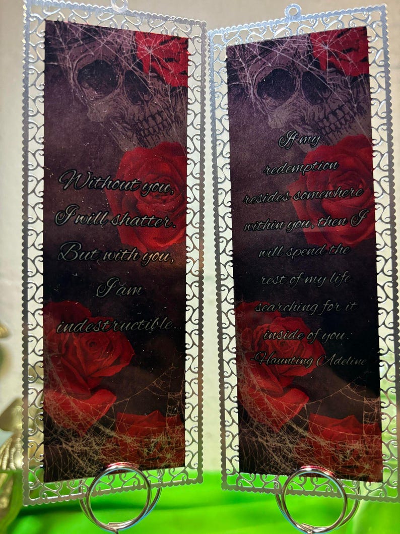 May include: Two silver bookmark holders with a skull and red roses design. The text on the bookmarks reads: "Without you, I will shatter. But with you, I am indestructible." and "If any redemption resides somewhere within you, then I will spend the rest of my life searching for it inside of you. Haunting Adeline."