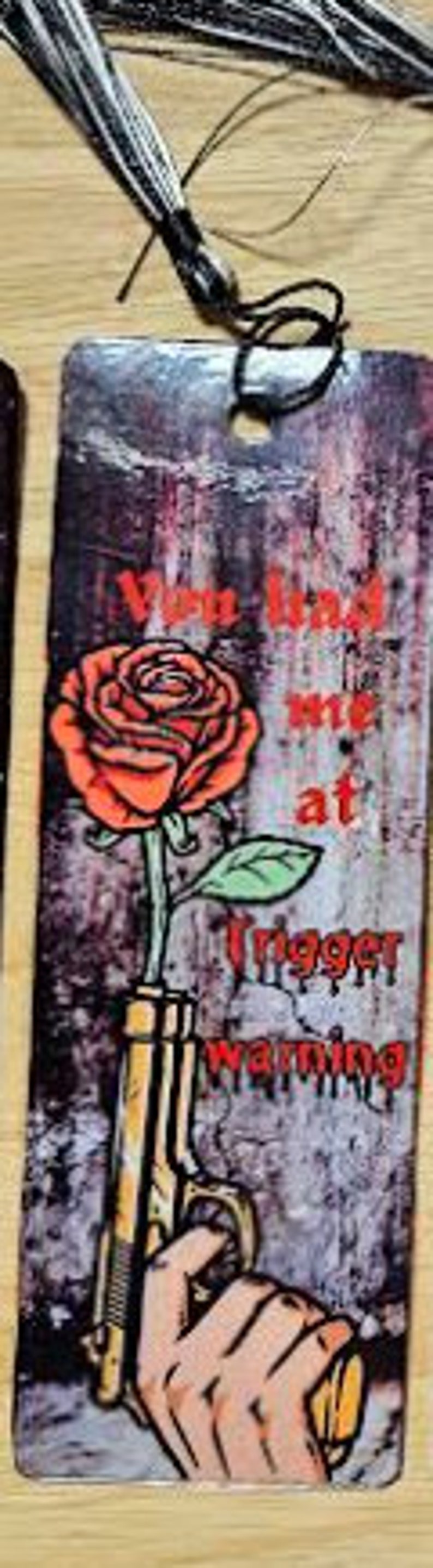May include: A bookmark with a grunge background featuring a red rose with a gun behind it. The text reads "You find me at Trigger Warning".