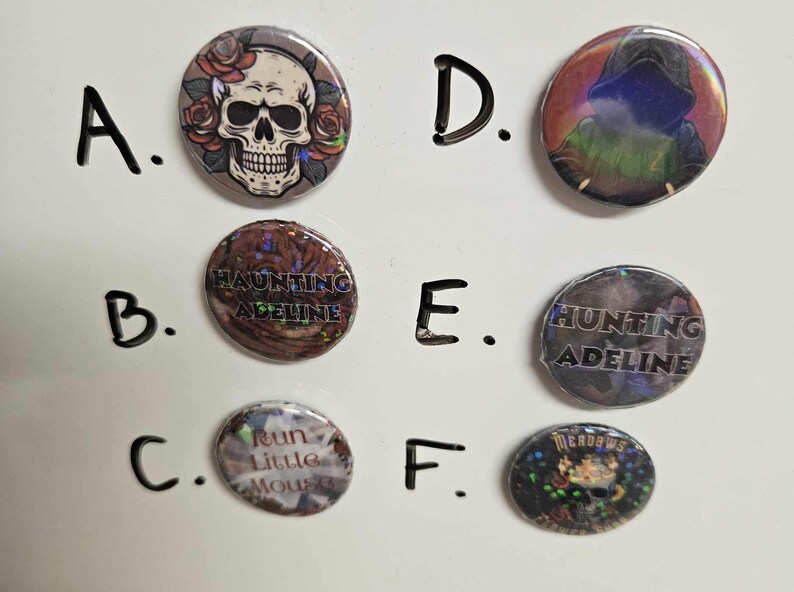 Puede incluir: Seis botones diferentes con varios dise&ntilde;os. Los botones son redondos y tienen un acabado brillante e iridiscente. Los dise&ntilde;os incluyen una calavera con flores, un dise&ntilde;o de texto que dice "Haunting Adeline", un dise&ntilde;o de texto que dice "Hunting Adeline", un dise&ntilde;o de texto que dice "Run Little Mouse", un dise&ntilde;o con una figura encapuchada y un dise&ntilde;o con una calavera y flores.