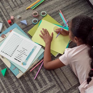 May include: A young person is drawing on yellow paper with a purple pencil. The scene includes colorful pencils, washi tape, and geometric shapes. A checklist with the word "AFTERNOON" is visible.