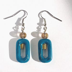 May include: A pair of turquoise blue dangle earrings with a silver hook and a brown bead. The earrings feature a rectangular shape with a smaller rectangle cut out in the centre.