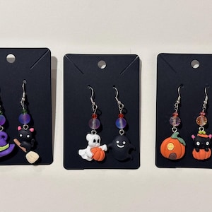 May include: Three pairs of Halloween-themed dangle earrings. Each pair features a different charm: a witch hat and black cat, a ghost and a black ghost, and a pumpkin and a black cat. The earrings have colorful beads and silver hooks.