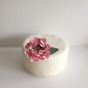May include: A single pink peony flower with a green leaf sits atop a white cake.