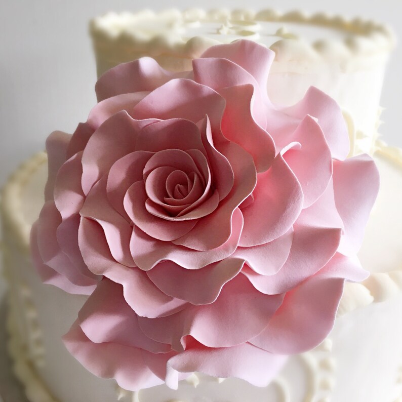 Op de afbeelding: Een roze fondantroos met gekrulde bloemblaadjes op een witte bruidstaart.