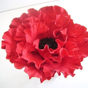 May include: A single red poppy flower with a black center. The flower is made of fabric and has a ruffled texture.