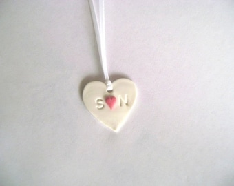 Op de afbeelding: Witte keramische hartvormige hanger met een roze hart en de letters "S" en "N" op een wit lint.