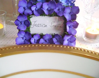 Op de afbeelding: Een klein wit frame met een paarse bloemenrand en de naam "Jessica Smith" geschreven in zwarte inkt. Het frame staat op een witte tafel met een gouden rand.