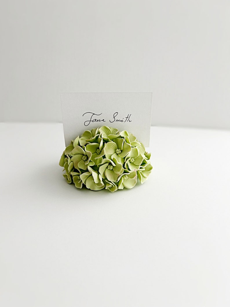 Puede incluir: Un porta tarjetas de lugar de flores de hortensia verde con una tarjeta blanca que dice "Jane Smith".