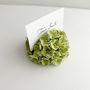 Puede incluir: Un arreglo floral de hortensia verde con una tarjeta blanca que dice "Jane Smith" para una tarjeta de lugar de boda o evento.