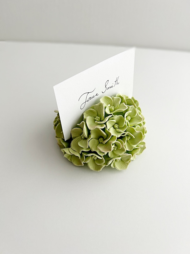 Puede incluir: Un arreglo floral de hortensia verde con una tarjeta blanca que dice "Jane Smith" para una tarjeta de lugar de boda o evento.