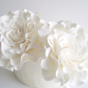 Puede incluir: Dos grandes rosas blancas de fondant con p&eacute;talos rizados, dispuestas sobre un pastel blanco.