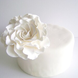 Puede incluir: Un pastel blanco con una gran rosa blanca de fondant en la parte superior.
